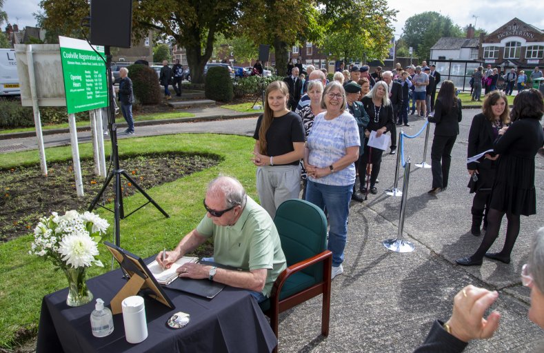 A man signs a book of condolence on a table with a photo frame and flowers, in front of a queue of people.