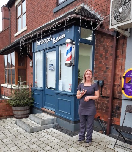 A lady dressed in a hairdressing outfit stands in front of Barthorpe's Barbers holding a pair of scissors