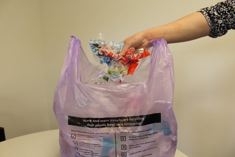 A picture of a hand placing a range of flexible plastic - carrier bags and packets - into a purple bin liner. These purple sacks will be used to collect flexible plastic from households on a trial.