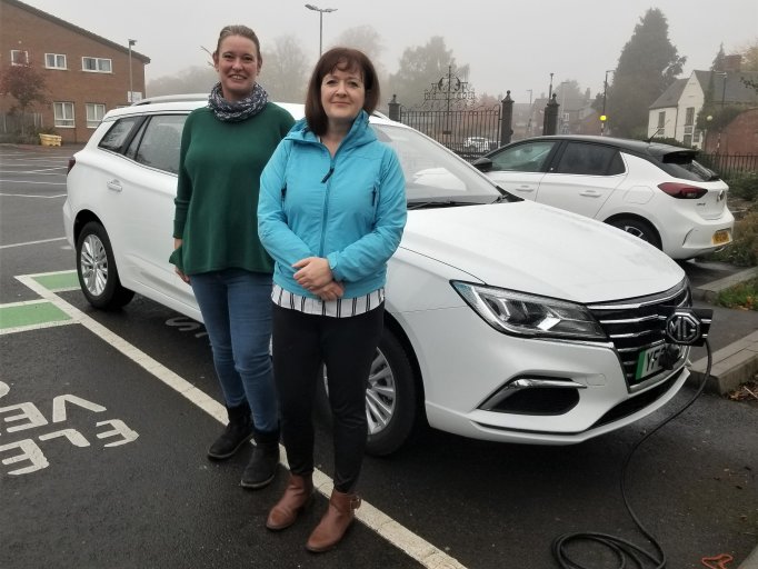 Two women standing in a car park next to a white estate MG car. The car is plugged into an electric charging point.