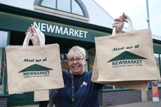 Image of woman standing outside Newmarket holding two shopping bags that say 'Meet me at Newmarket', with the market sign in the background.
